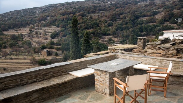 An outdoor patio with stone flooring, a stone table, and wooden chairs. - Petra Traditional House (Andros)
