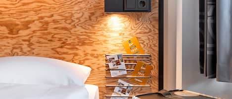Habitación de hotel con cabecera de madera, cama con ropa blanca, caja de almacenamiento negra con un logotipo e iluminación fijada a la pared.