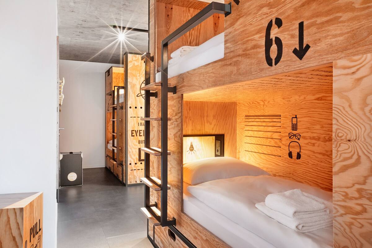 A modern, compact room with a bunk bed, wooden walls, and a pull-down desk.