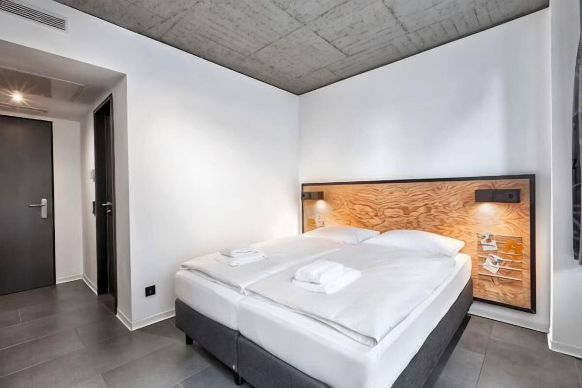 A modern hotel room with a large bed, a wooden headboard, and a wall-mounted light fixture.