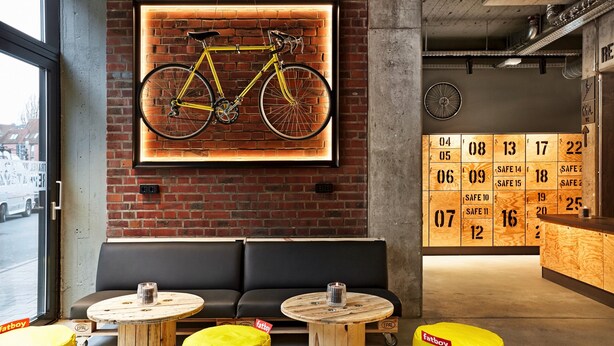 Un interior moderno con una bicicleta colgada en una pared de ladrillo, un sofá negro y mesas redondas de madera con taburetes amarillos.