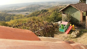Une cabane rustique avec un toit végétalisé, un petit espace de jeux avec un toboggan, et une vue sur des collines ondoyantes et une vallée en arrière-plan.