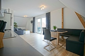 En moderne lejlighed med køkken, spiseområde og opholdsstue, med et stort vindue, en plante og et fjernsyn.