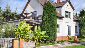 Een twee verdiepingen tellend huis met een balkon, een hoge naaldboom en een bakstenen muur.