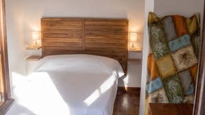 A bedroom with a wooden headboard, two bedside lamps, a bed with white bedding, and a colorful patterned throw on a wooden chair.