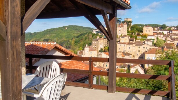 A balcony with a view of a village, a wooden pergola, and a white plastic chair.