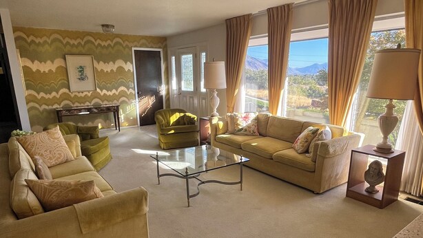 Una sala de estar con un sofá color beige, un sillón verde, una mesa de centro de vidrio y vistas al exterior a través de amplios ventanales.