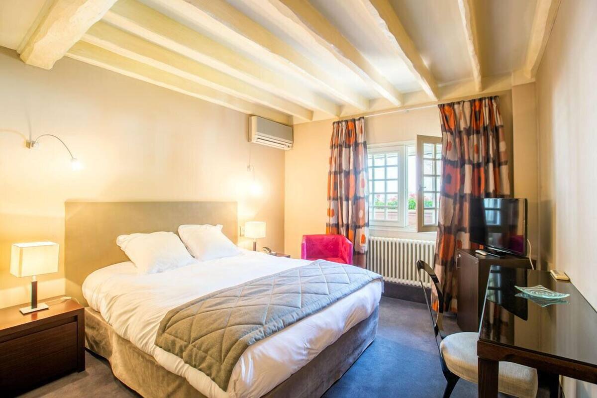 A hotel room with a bed, a desk, a chair, a TV, a window with curtains, and a radiator.