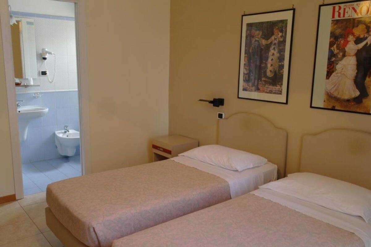 Quarto de hotel com duas camas de solteiro, uma mesa de cabeceira, um criado-mudo pequeno, uma televisão montada na parede e um banheiro visível através de uma porta aberta.
