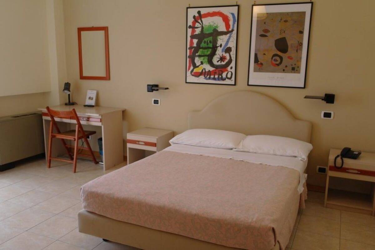Quarto de hotel com cama, escrivaninha, cadeira e duas obras de arte emolduradas na parede.