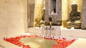 Una bañera llena de agua y pétalos de rosa, con estatuas de piedra al fondo.