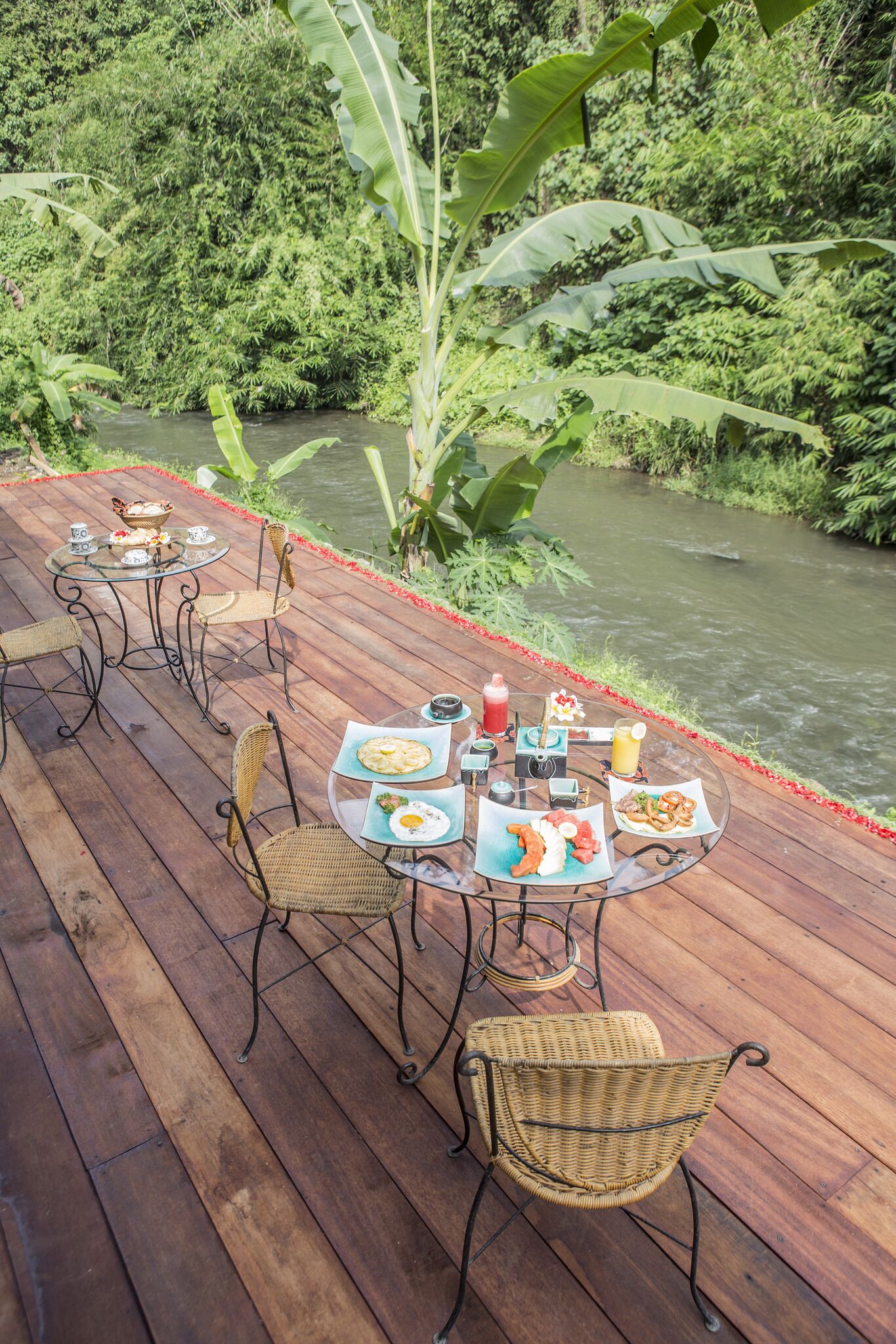 A wooden deck with round tables and wicker chairs, overlooking a river and lush greenery.