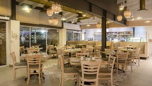 A restaurant interior with multiple dining tables and chairs, a bar area, and a view of the city through large windows.