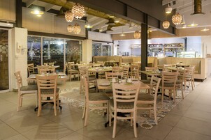 Interior de restaurante con varias mesas y sillas, una zona de bar y vistas a la ciudad a través de amplios ventanales.