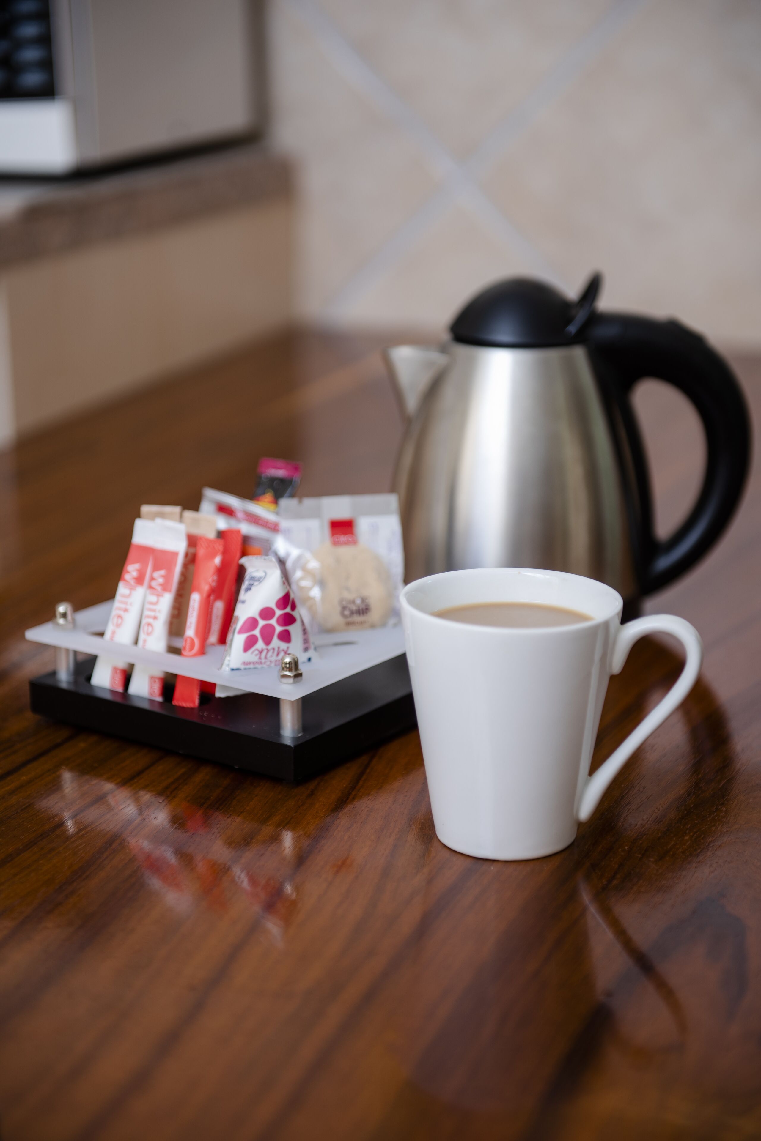 Una taza blanca sobre una mesa de madera, con una bandeja de alimentos envasados y una tetera de acero inoxidable al fondo.
