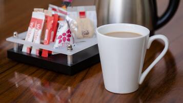 Una taza blanca sobre una mesa de madera, con una bandeja de alimentos envasados y una tetera de acero inoxidable al fondo.
