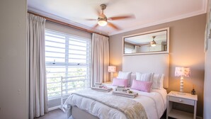 Un dormitorio con una cama, dos mesitas de noche, un ventilador de techo, una ventana con cortinas y un espejo en la pared.