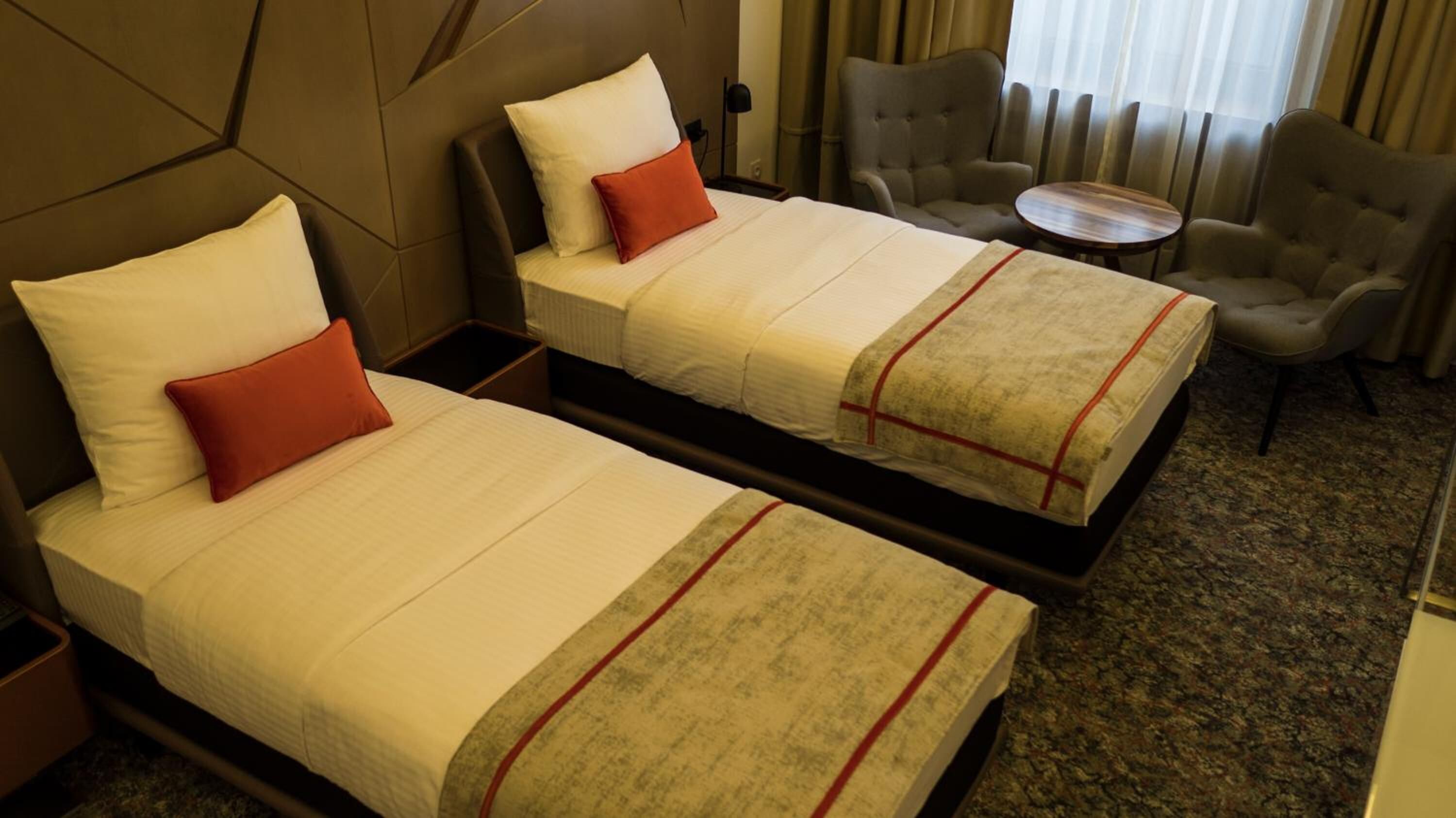 Ein Hotelzimmer mit zwei Betten, jeweils mit Kissen und Decke. Zwei Sessel und ein kleiner Tisch sind ebenfalls vorhanden.