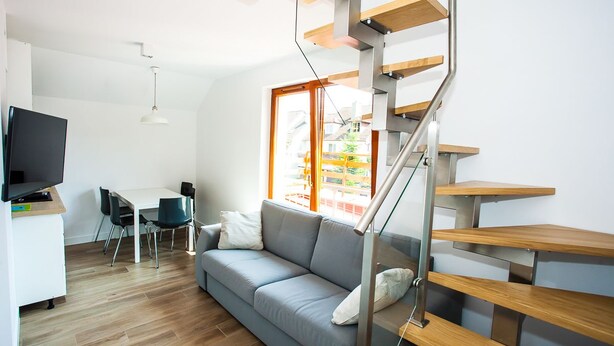 A modern living space with a grey sofa, a dining table with chairs, a TV, and a staircase with wooden steps. - Apartamenty SaltApart 2 (Wieliczka)