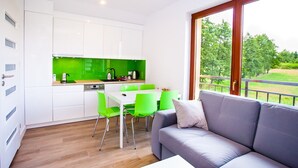 A modern kitchen with white cabinets and a green backsplash, a dining area with green chairs, and a grey sofa with a view of a green landscape through large windows. - Apartamenty SaltApart 2 (Wieliczka)