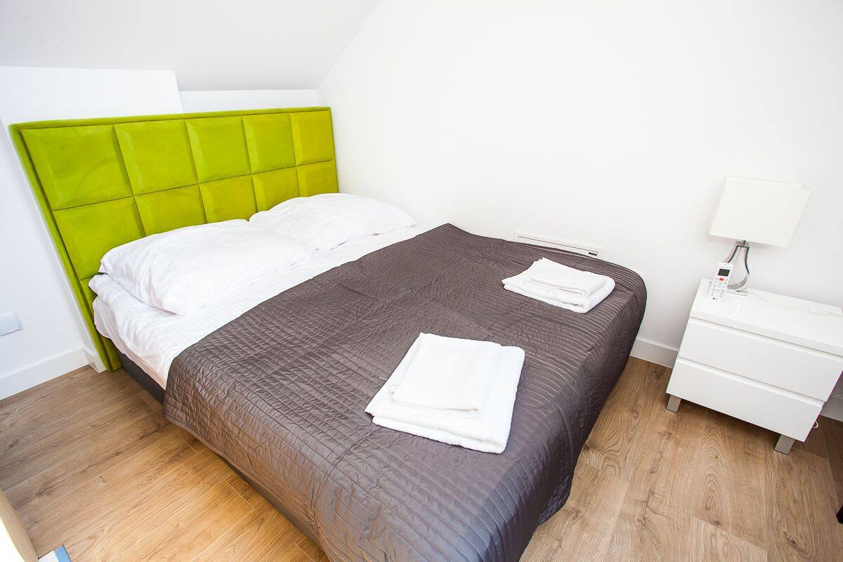 A neatly made bed with a green headboard, a white bedside table with a lamp, and a wooden floor.
