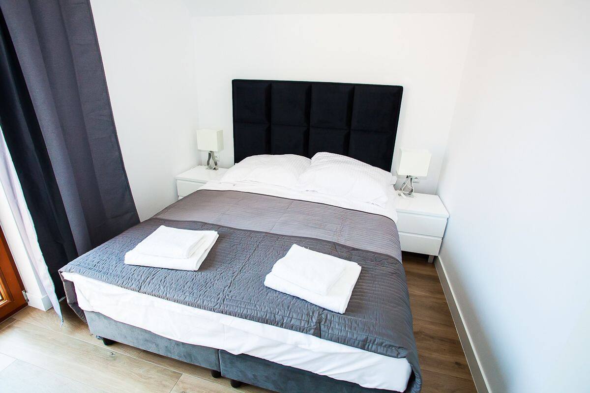 A neatly made bed with a grey and white duvet, two white towels on the bed, a black headboard, and a white bedside table with a lamp.