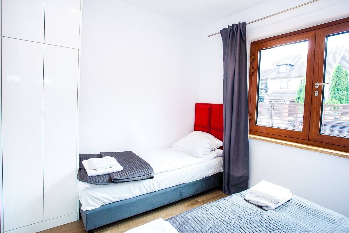 A small, modern bedroom with a single bed, a red headboard, and a window with curtains.