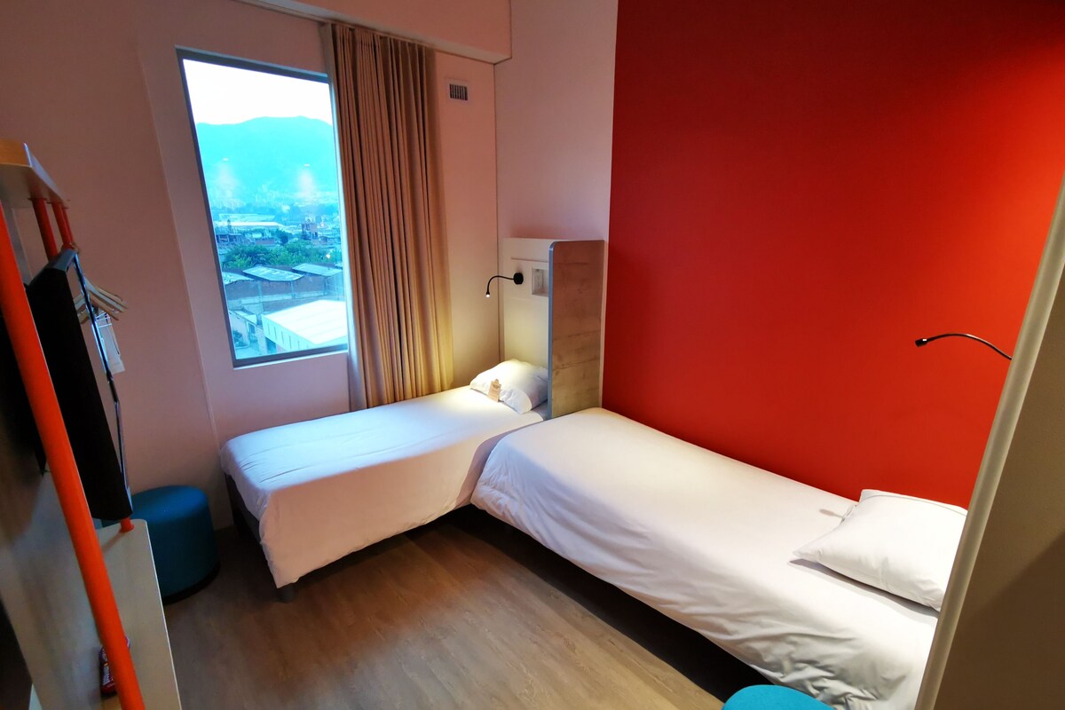 A hotel room with two beds, a large window with a view, a red accent wall, and a blue chair.
