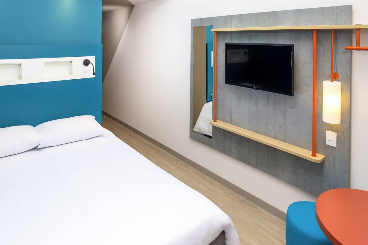 A hotel room with a bed, a TV mounted on the wall, a small table, and a chair.