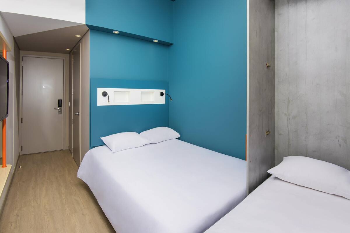 A hotel room with a single bed, a bedside table, a TV mounted on the wall, and a door leading to another room.