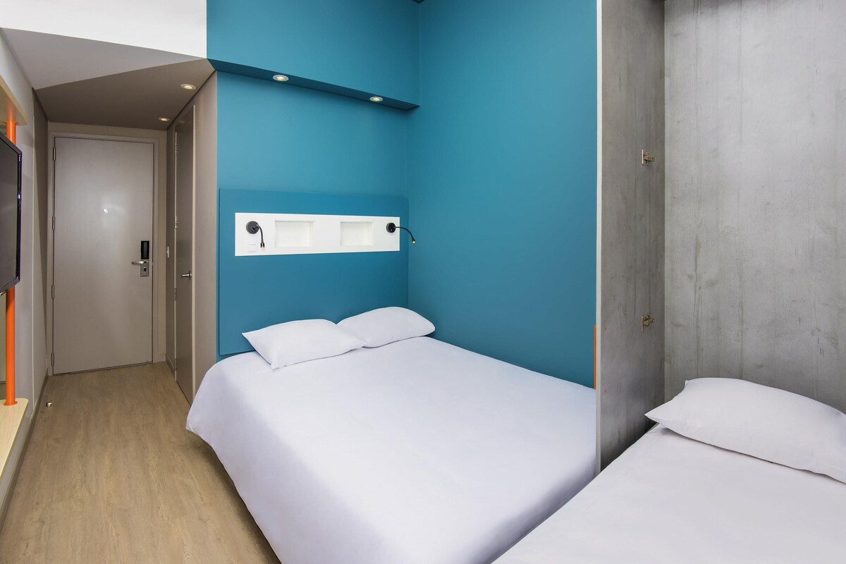 A hotel room with a single bed, a bedside table, a TV mounted on the wall, and a door leading to another room.