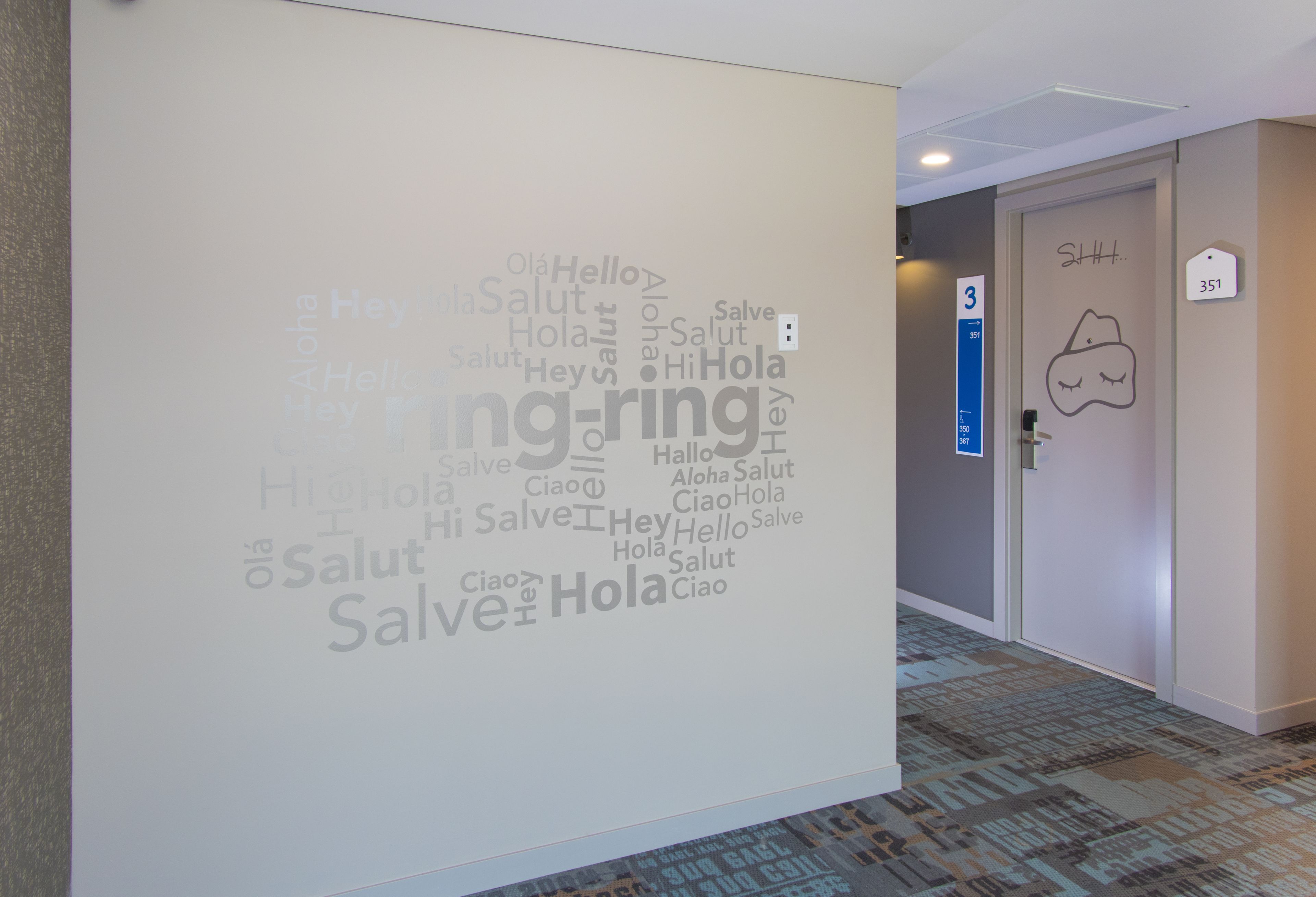 Un couloir avec un mur orné d’un nuage de mots contenant des salutations dans différentes langues, une porte portant l’inscription « 3 » et une autre porte avec le numéro « 351 ».