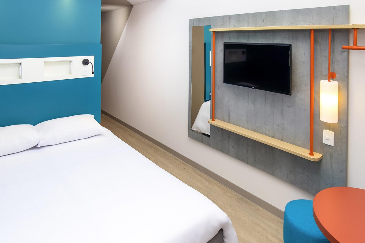 A hotel room with a bed, a TV mounted on the wall, a small table, and a chair.