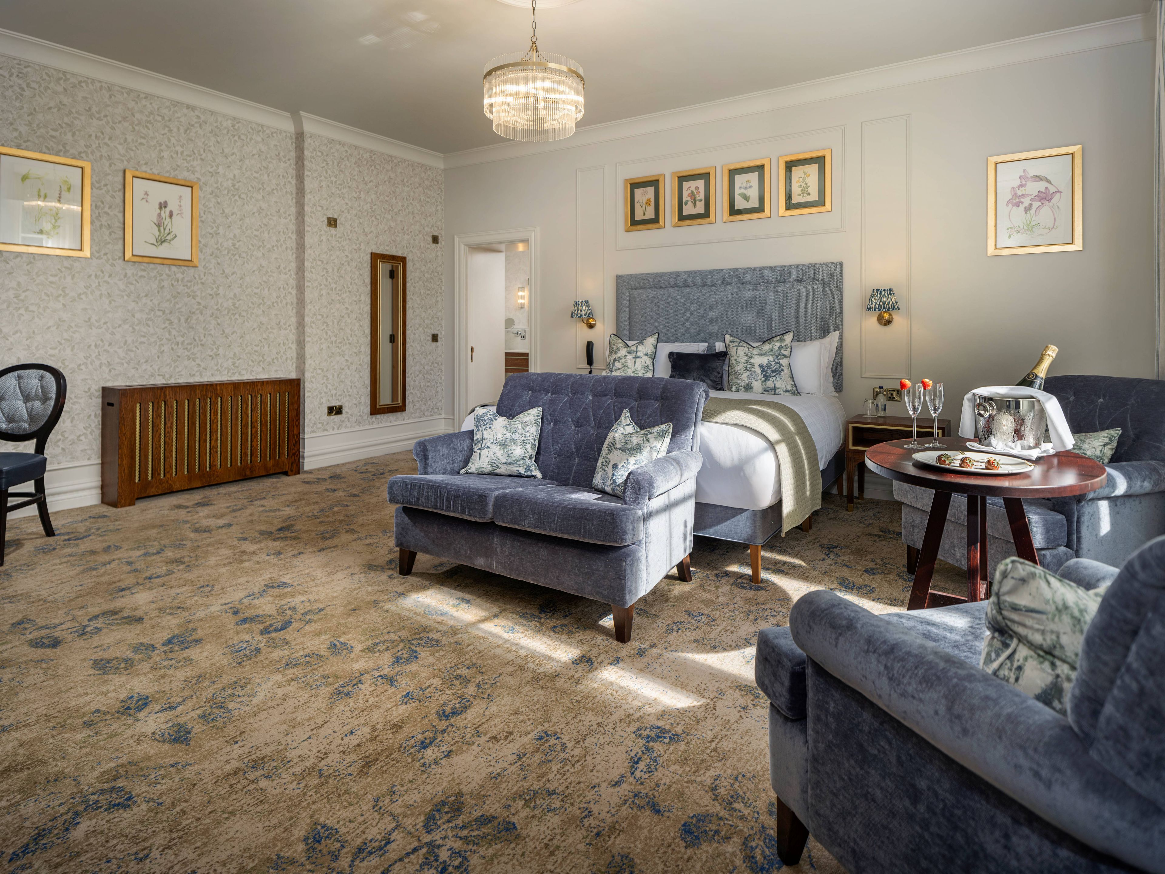 A hotel room with a large bed, two armchairs, a small table with a bottle of champagne, and framed botanical prints on the walls.