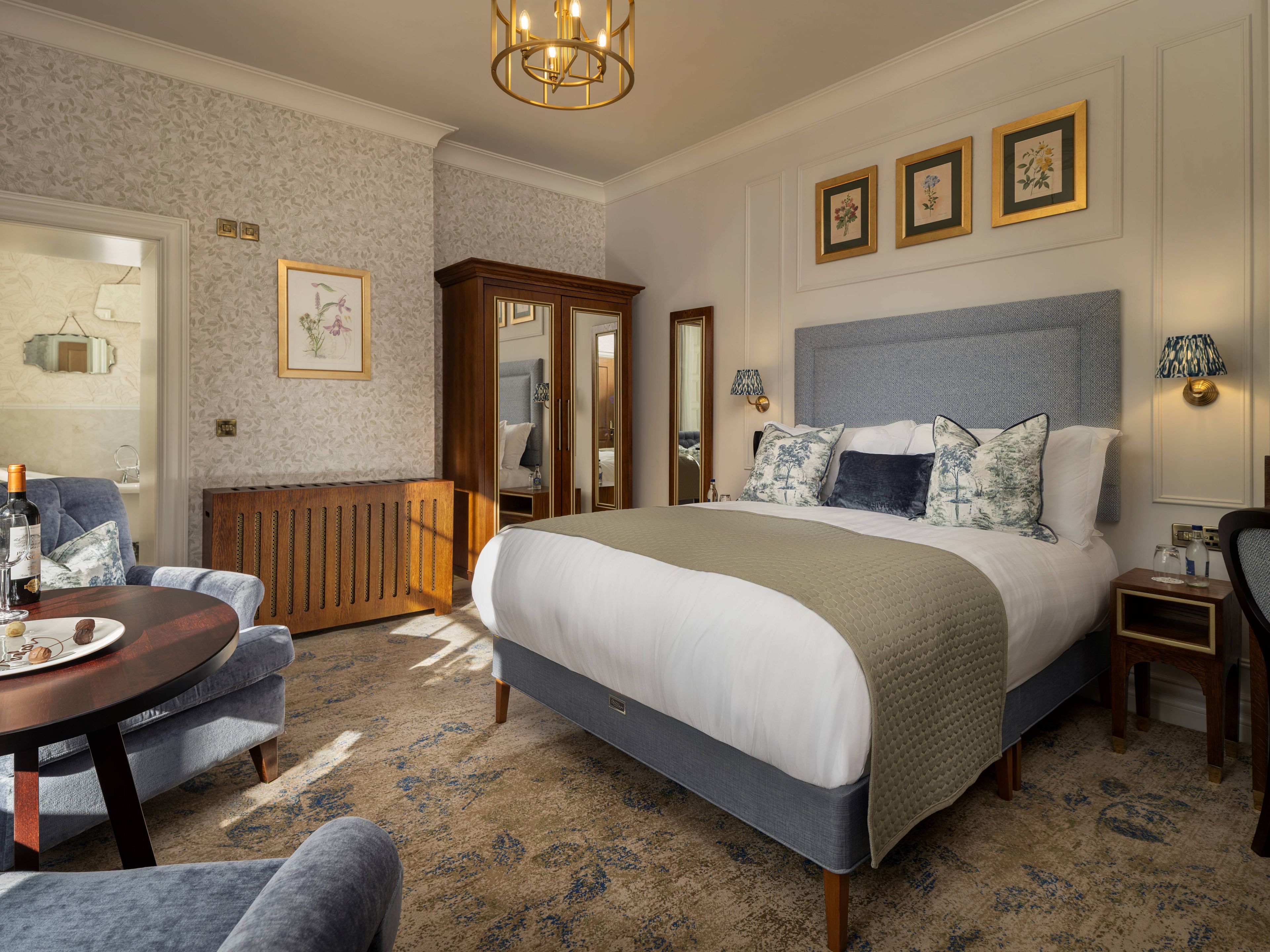 A hotel room with a large bed, a wooden cabinet, a round table with a bottle, and a chair.