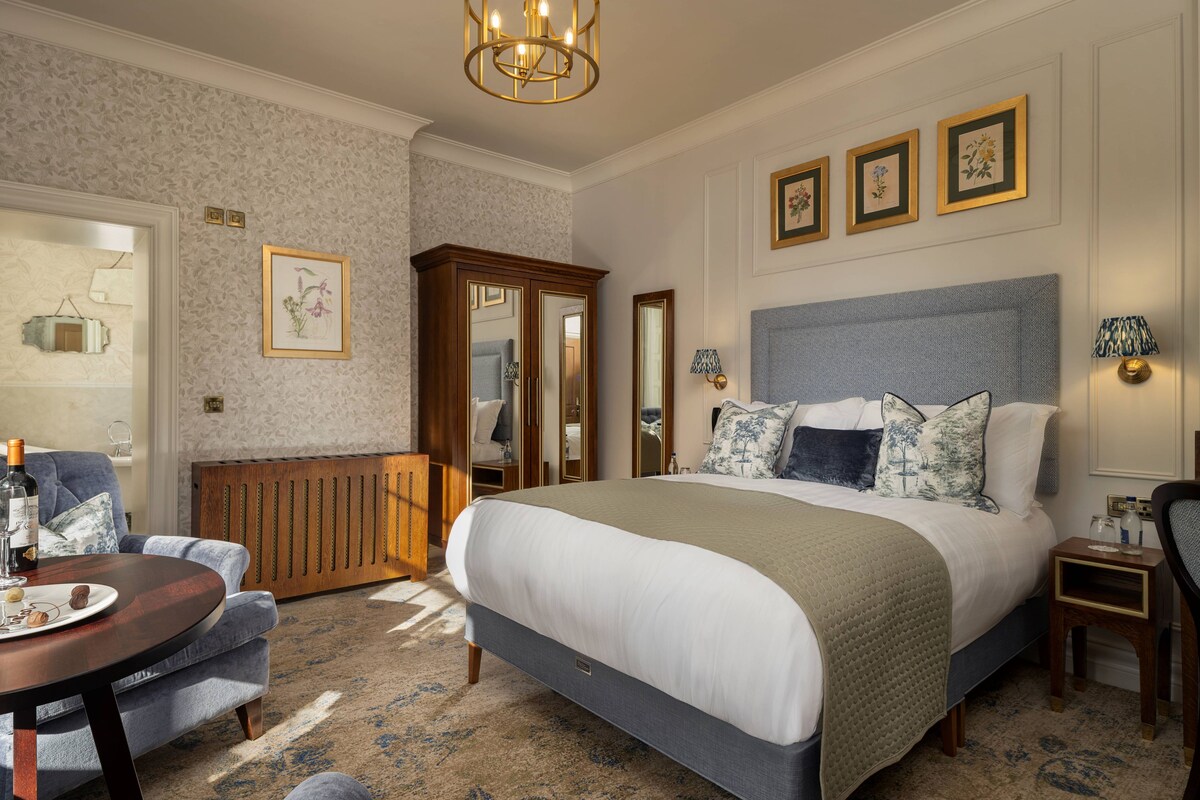 Una habitación de hotel con una cama grande, un armario de madera, una mesa redonda con una botella y una silla.