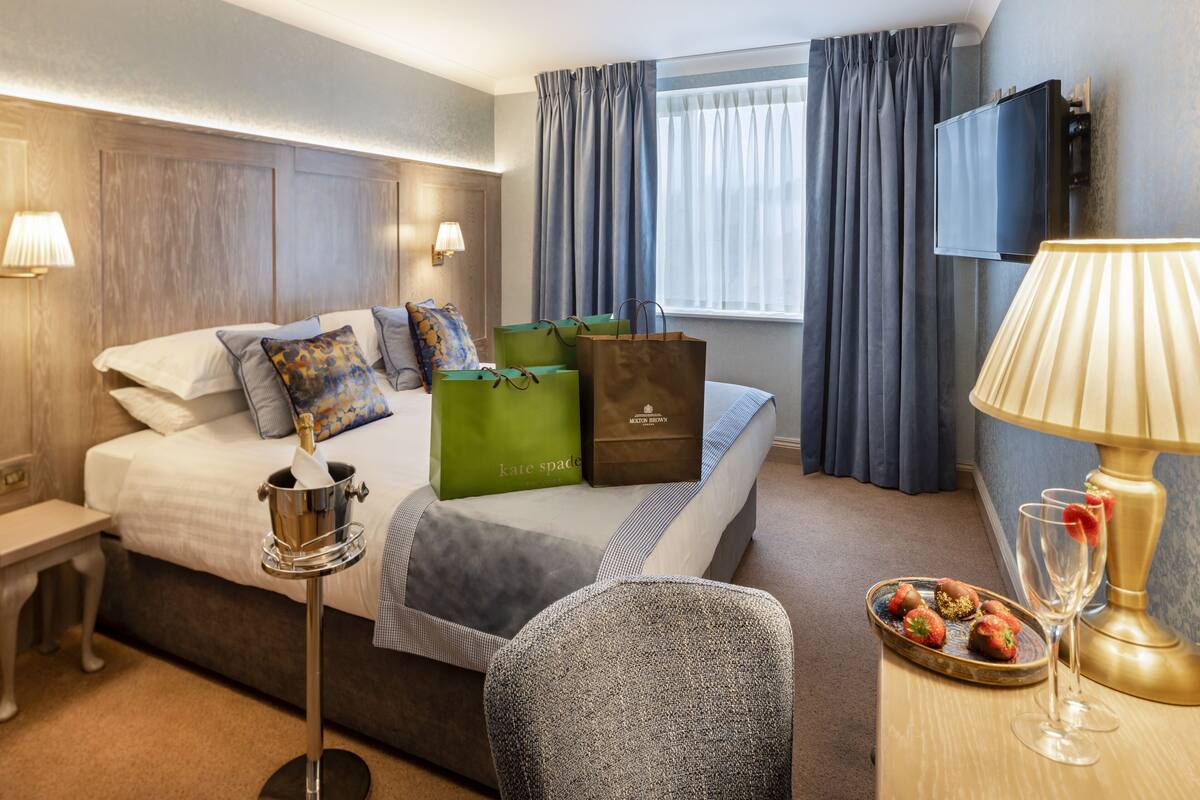 Habitación de hotel con cama, almohadas, mesita de noche con lámpara, una silla, bandeja con fresas y ventana con cortinas.