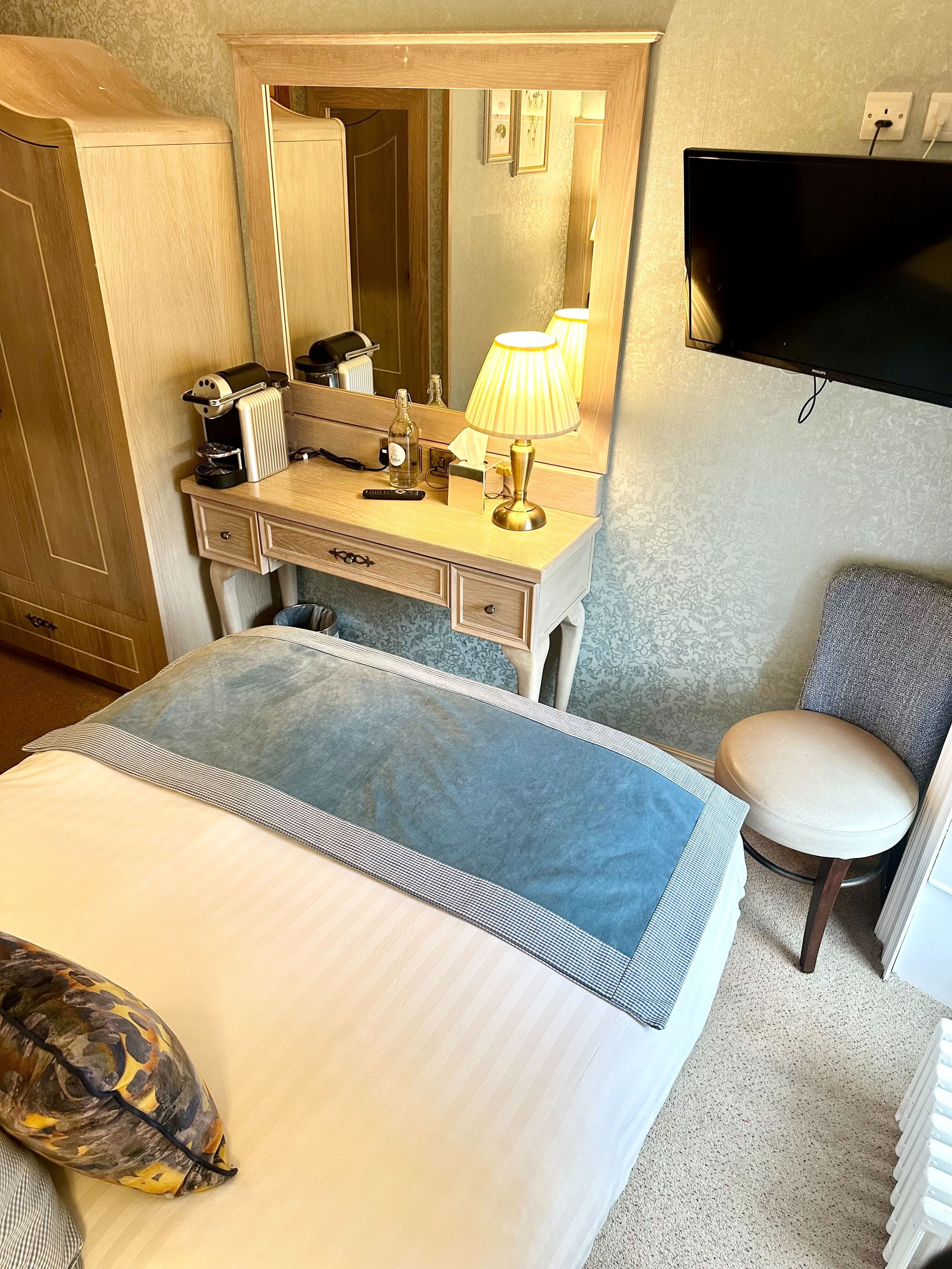 Habitación de hotel con cama, escritorio con lámpara, silla, televisor y radiador.