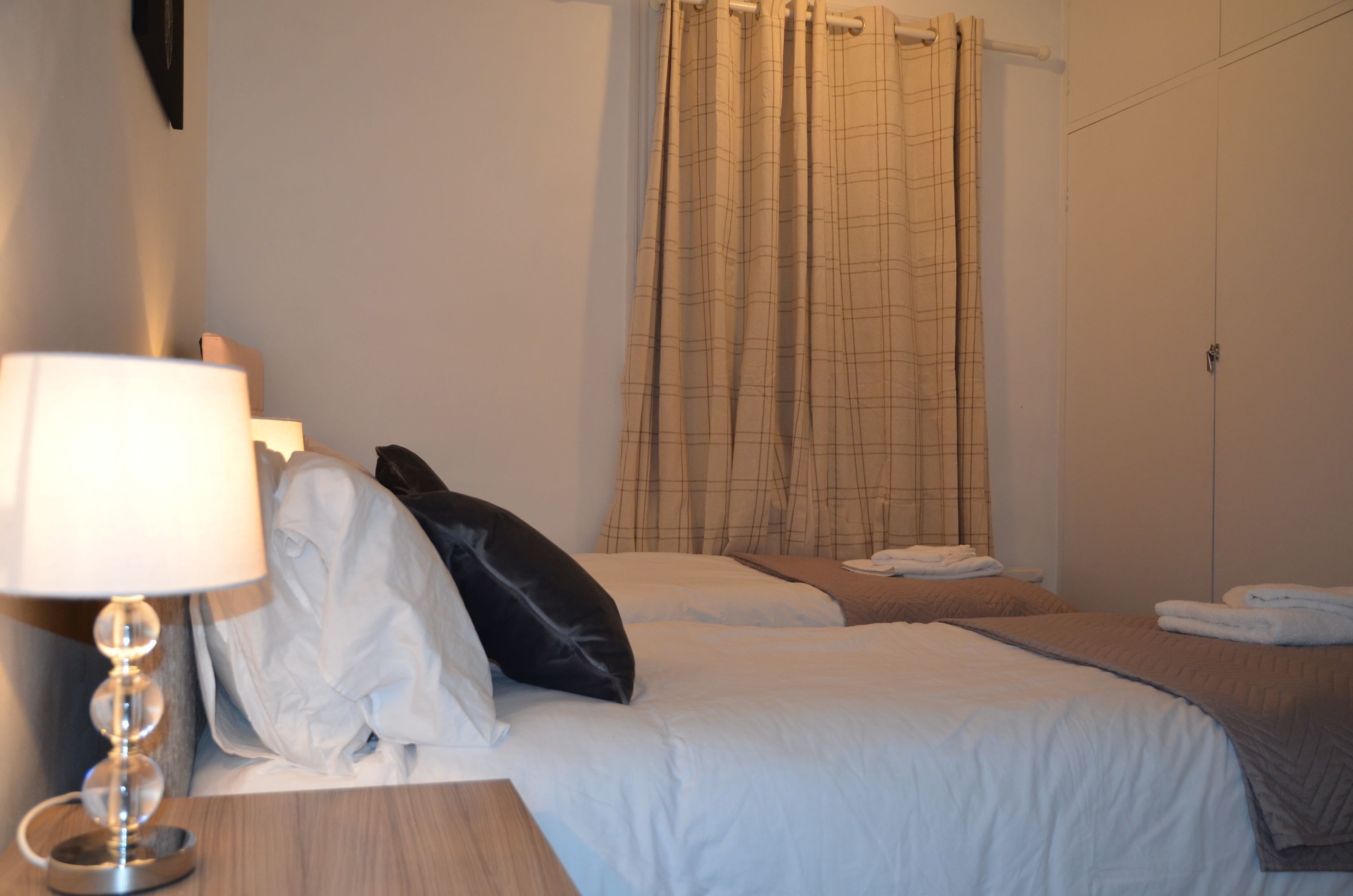 A hotel room with two beds, a bedside table with a lamp, a curtain, and a wardrobe.