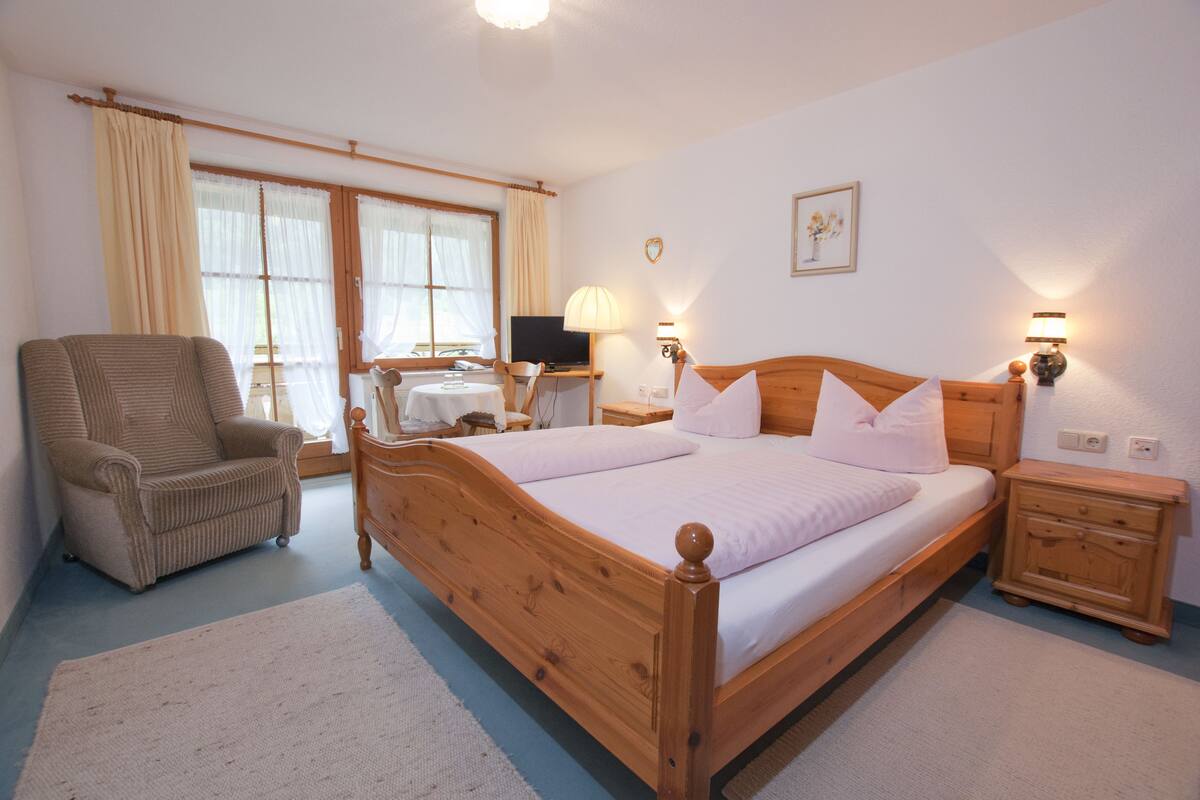 Quarto com cama de madeira, uma cadeira, escrivaninha com computador e janela com cortinas.