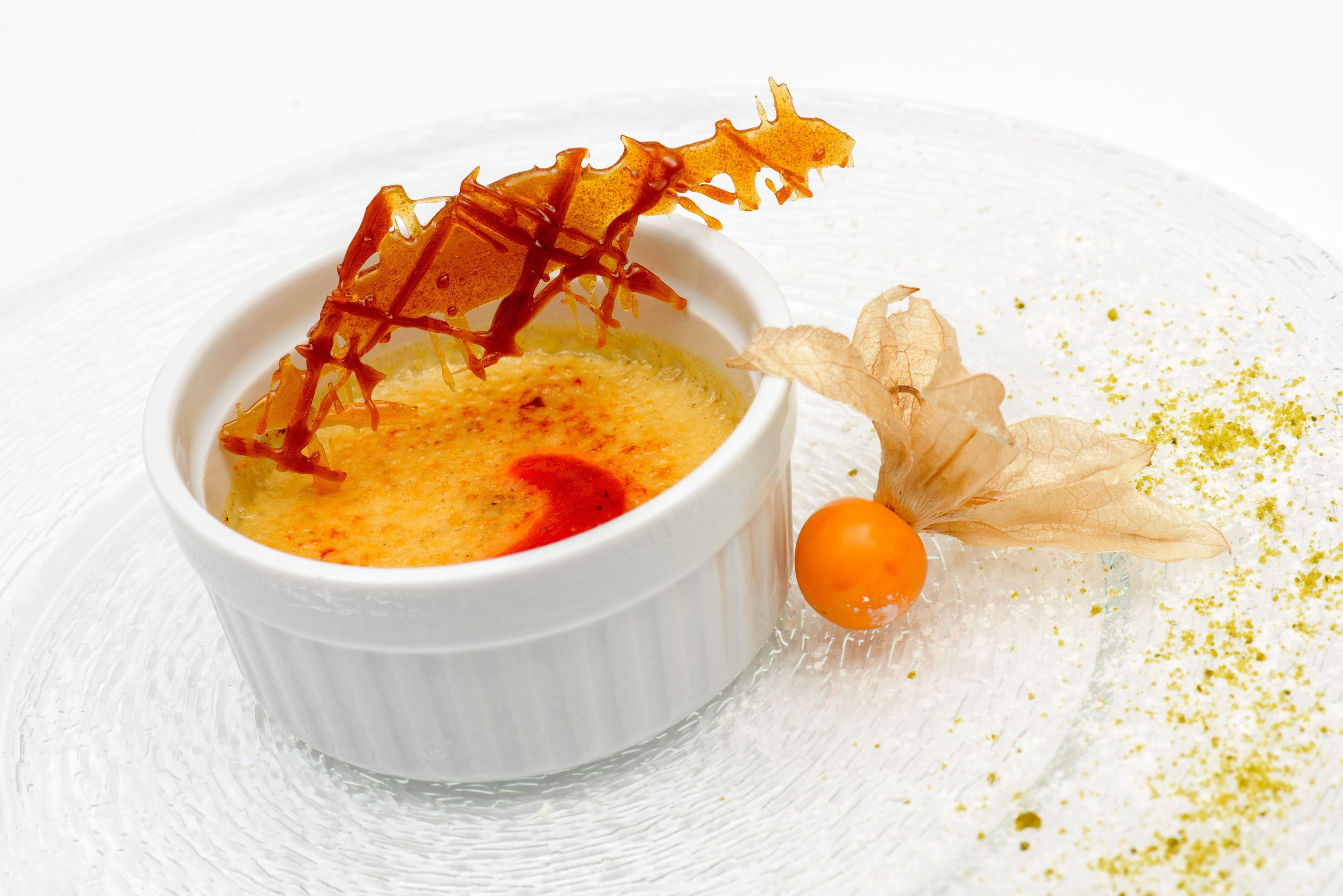 A dessert in a white bowl with caramelized sugar and a decorative leaf on a white textured surface.