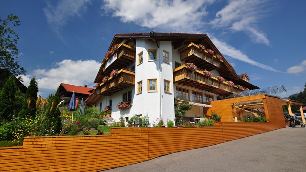 A multi-story building with balconies adorned with flowers, a wooden fence in front, and a clear blue sky. - Berghüs Schratt - vegetarisches und veganes Biohotel (Oberstaufen)