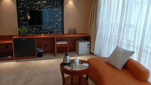Habitación de hotel con sofá, una mesita con tetera, televisión, pared de mármol, ventana con cortinas y minibar.
