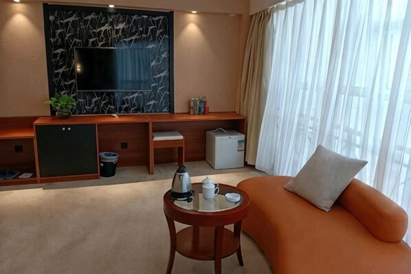 A hotel room with a sofa, a small table with a kettle, a TV, a marble wall, a window with curtains, and a minibar.