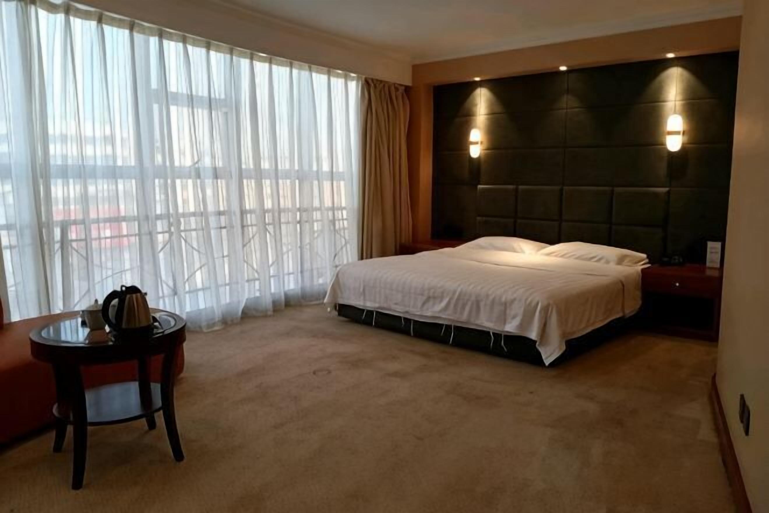 A hotel room with a large bed, a bedside table, a chair, a window with curtains, and a headboard with wall-mounted lights.