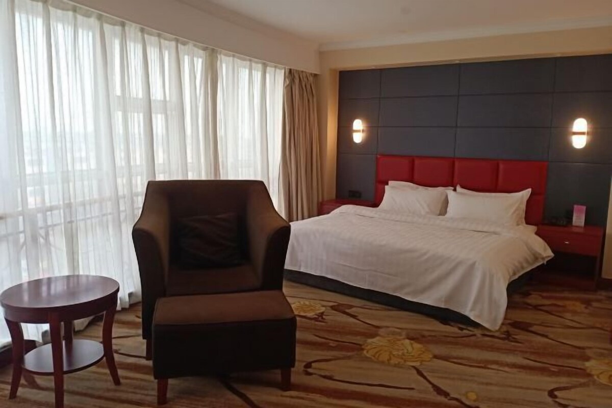 Una habitación de hotel con cama, una silla, una mesita pequeña y un ventanal con cortinas.