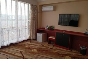 Una habitación con alfombra estampada, un refrigerador blanco pequeño, un banco rojo, un televisor montado en la pared y una ventana con cortinas traslúcidas.