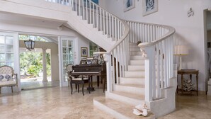 A grand staircase with white balustrades, a piano, and a dining area with a table and chairs. - Whispering Palms (Belair)