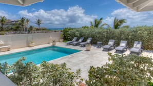 A swimming pool with lounge chairs, surrounded by greenery and a view of the ocean. - Whispering Palms (Belair)