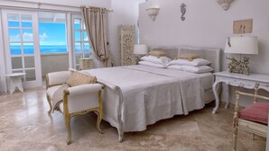 A spacious bedroom with a large bed, two chairs, a nightstand, and a view of the sea through a large window. - Whispering Palms (Belair)